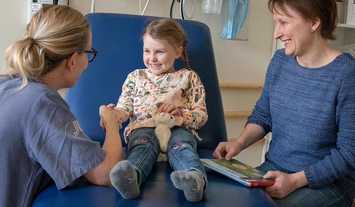 Ung flicka som sitter på en brist och håller en kvinna med sjukhuskläder i handen och en kvinna som håller i en bok