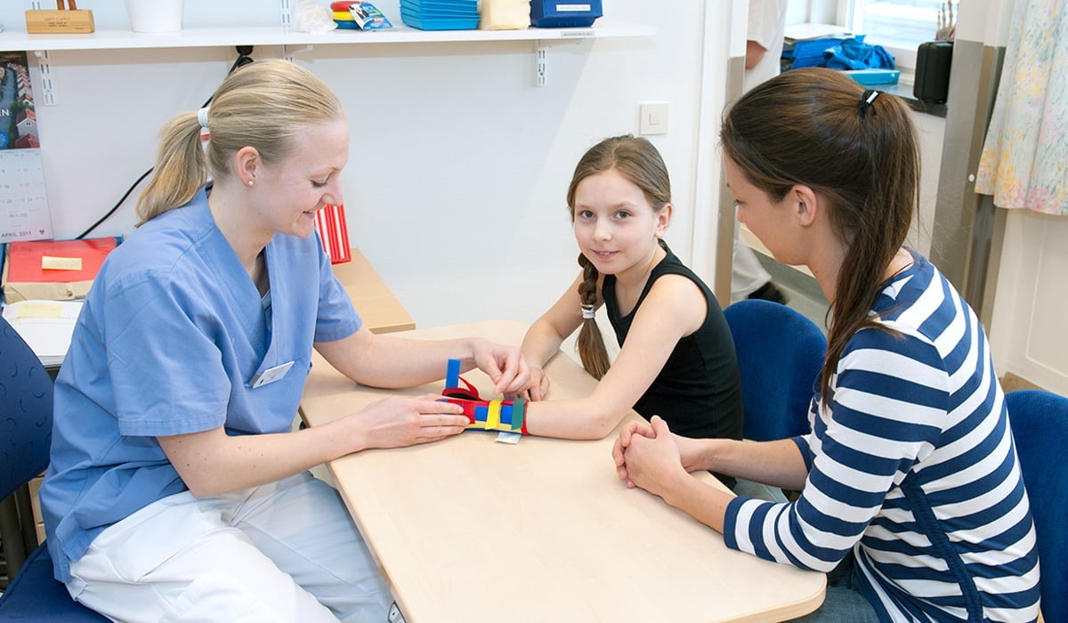 Bild på en kvinna med sjukhuskläder som hjälper en ung flicka med en skena på handen. En till kvinna i privata kläder sitter bredvid unga flicka
