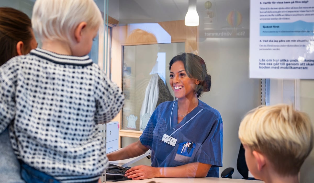 Kvinna med sjukhuskläder som sitter i en reception med två barn och en vuxen i bakgrunden
