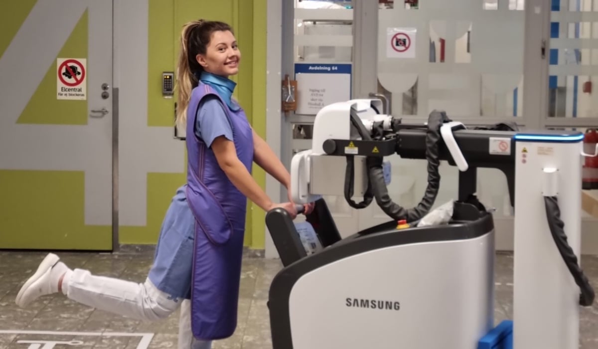 kvinnlig sjuksköterska skjuter röntgenmaskin framför sig i sjukhusmiljö.